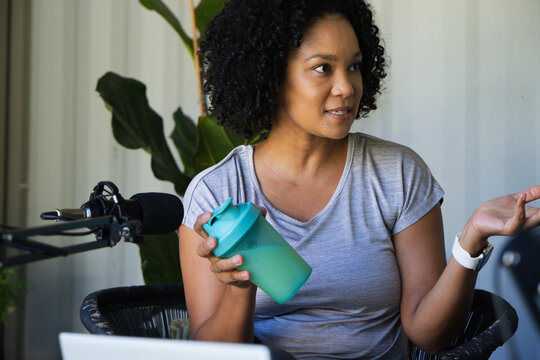 African American woman sitting in woven chair at home studio holding teal cup, gesturing to mic
