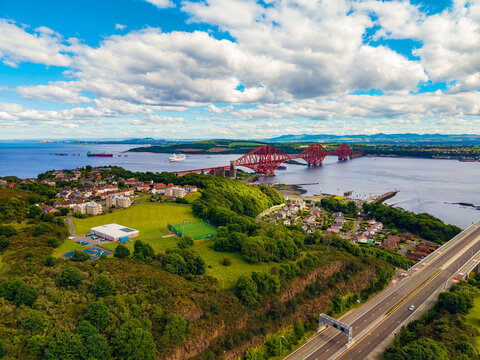 Drone view of the red Forth Bridge, a UNESCO rail landmark, spanning the Firth of Forth by North Queensferry near Edinburgh, Scotland, with green coast and clouds.