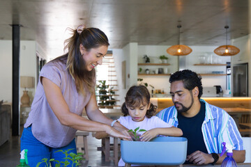 Diverse family guiding child's hands planting seedling in planter on wooden table in modern kitchen