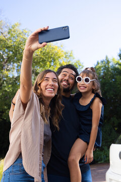 Diverse family taking selfie on driveway, woman holding smartphone, child wearing white sunglasses