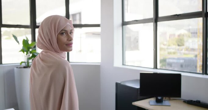 African American woman wearing hijab turning and smiling by window desk plant, responding to camera
