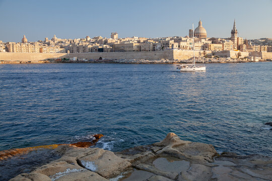 A sailboat drifts across calm blue harbor waters with Valletta historic limestone skyline, domes and spires bathed in warm sunset light, evoking coastal serenity of Malta, travel and architecture