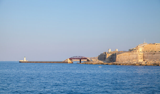 A stone fortress and lighthouse sit along a long pier and breakwater as a small boat drifts on calm blue water under clear evening sky. Valletta, Malta