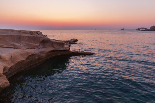 Soft pastel sunset light bathes a rocky coastline and calm sea, creating a tranquil, reflective mood perfect for travel, nature, and coastal lifestyle visuals at dusk. Sliema, Malta