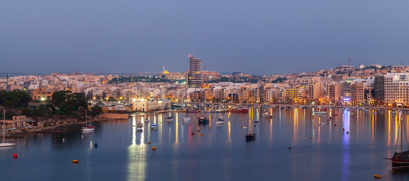 Calm Mediterranean harbor at dusk with moored boats and yachts, illuminated waterfront buildings and colorful light reflections on the water conveying evening tranquility and coastal urban life. Malta