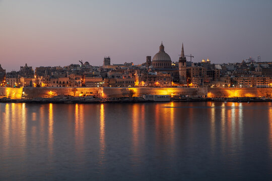 Historic Valletta skyline illuminated at dusk, warm city lights reflecting on calm Maltese harbor waters. Atmospheric Maltese architecture, domed cathedral and fortified waterfront at night