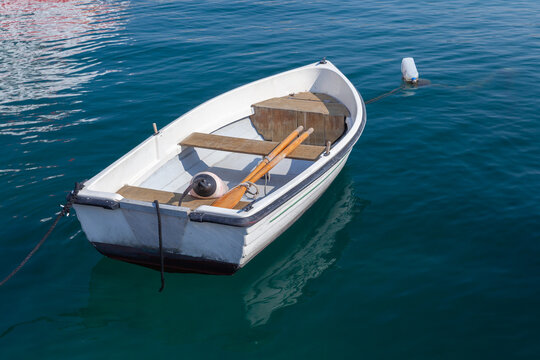 A small white rowboat moored to a buoy and floating on calm blue harbor water, with wooden oars resting inside. Peaceful, summery scene conveying leisure, boating, and coastal relaxation. Malta