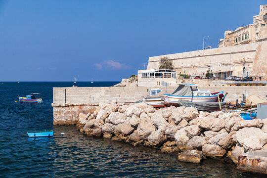 Colorful fishing boats moored by a rocky breakwater and stone quay, overlooking calm Mediterranean blue sea under a clear sky, evoking coastal life, summer warmth and seaside charm of Malta