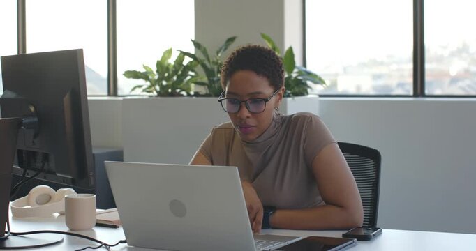 Glancing at laptop, African woman leaning in and typing on laptop at office desk, finishing report