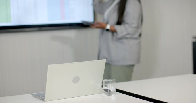 African woman in blazer entering right, pointing, explaining data on screen at table, copy space