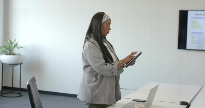 Adult African American woman holding tablet tapping and cross-checking display data in gray jacket