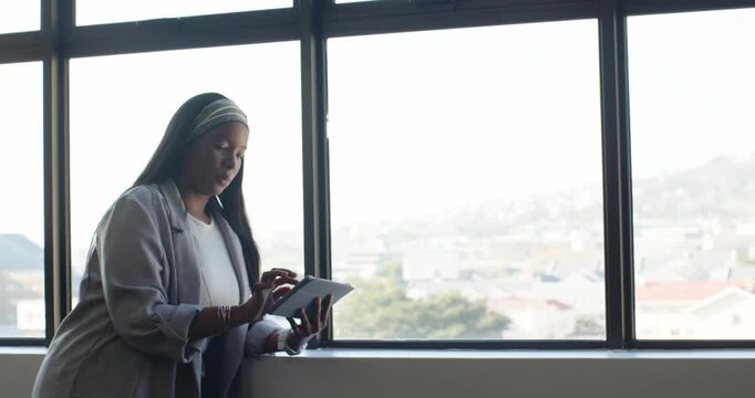 African adult woman leaning on window using tablet reviewing work wearing gray blazer, copy space
