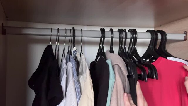Hands sorting and moving clothes on hangers in wardrobe, organizing garments at home, decluttering process, horizontal video