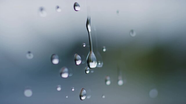 Slow motion close up of raindrops sliding and merging on window glass with blurred outdoor landscape background