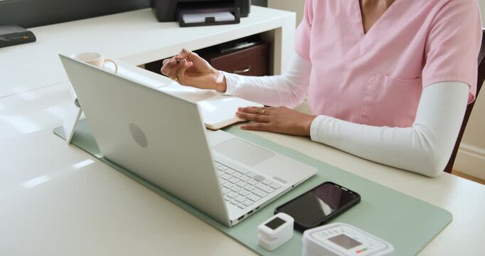 African American woman in pink scrubs at desk, laptop open, writing notes, typing cuff for e-visit