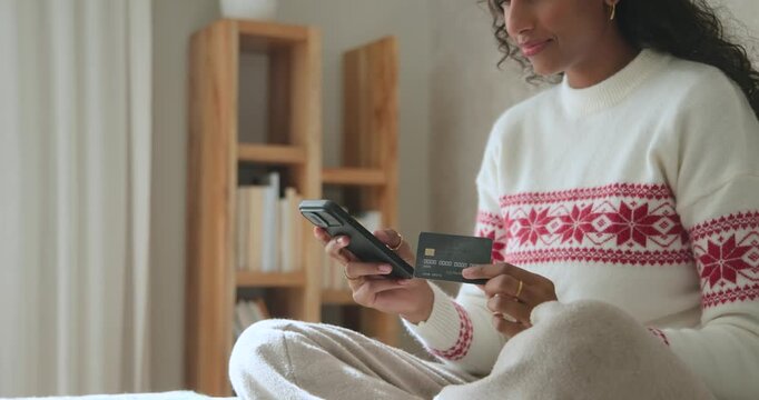 African-American woman in white sweater reading card, typing on phone, completing order in bed