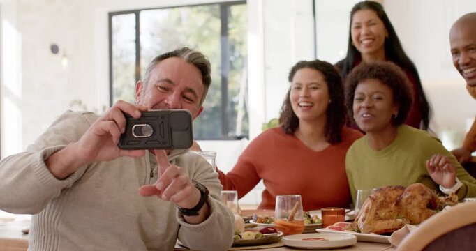 Diverse friends leaning in as smiling man holding black phone framing selfie at table with roast