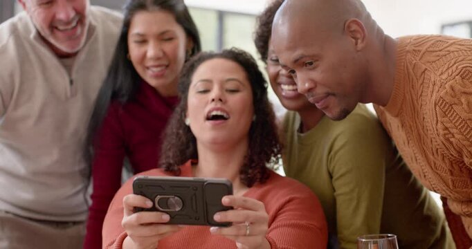 Diverse friends leaning in at home as seated woman extending smartphone case ring, sharing delight