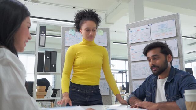 Confident businesswoman presenting technical charts and diagrams pinned to a wall, engaging with two colleagues seated at a desk in a modern tech office.