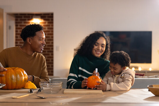 African American family carving pumpkins at dining table in sweaters, using spoon and carving tool