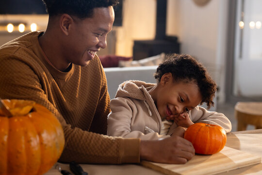 African American dad in brown sweater daughter in beige jacket carving pumpkins at home with tools