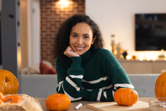 African American woman in green sweater resting chin on hand smiling, carving pumpkins at home