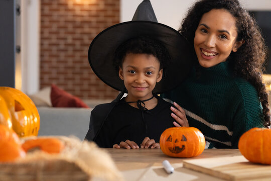 African American mom and child sitting carving pumpkins at home table witch hat, green sweater
