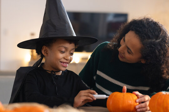 African American mother and daughter carving pumpkins at table, child in witch hat using small tool