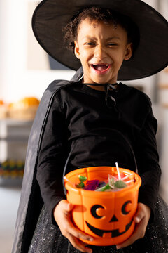 African American girl child in witch hat and black cape standing in kitchen holding pumpkin pail