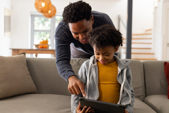 African American father and daughter leaning over gray sofa in living room, pointing at tablet