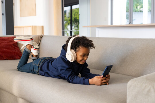 African American female child lying on sofa at home wearing headphones and holding smartphone