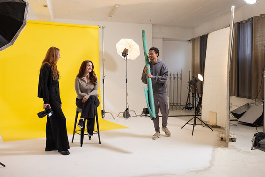 Diverse coworkers preparing studio shoot with yellow backdrop, DSLR camera, teal rolls and lights