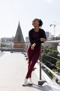 African American woman wearing maroon leggings white sneakers smiling, leaning on rail on roof