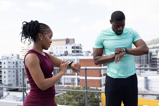 African couple standing on rooftop terrace in athletic wear checking smartwatches by metal railing