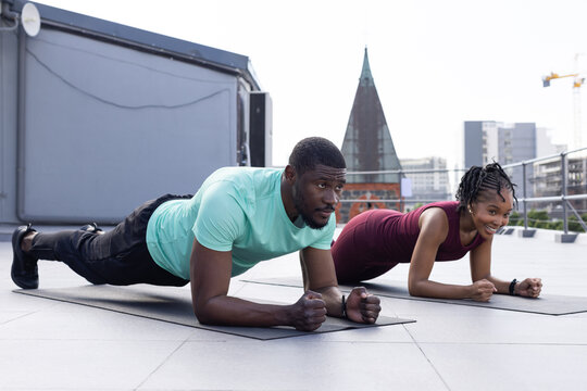 African American workout partners holding forearm planks in sportswear on rooftop with mats