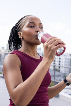 Adult African woman drinking from maroon bottle on roof, wearing maroon tank top and fitness watch