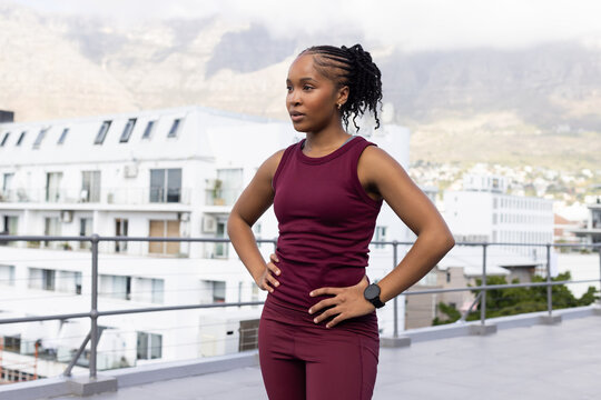 African woman standing on roof near metal railing in maroon gear, hands on hips checking smartwatch