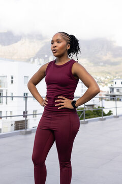 African American woman standing on rooftop in burgundy leggings pausing by railing with watch