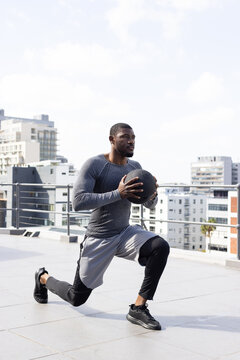 African American man lunging on rooftop holding black medicine ball, wearing compression leggings