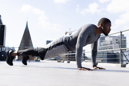 Mid-adult African American man performing plank on roof wearing long-sleeve top near railing