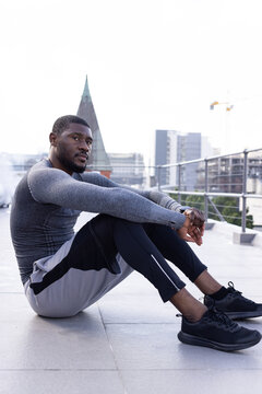 African American man sitting on rooftop by metal railing wearing tight gear and black shoes