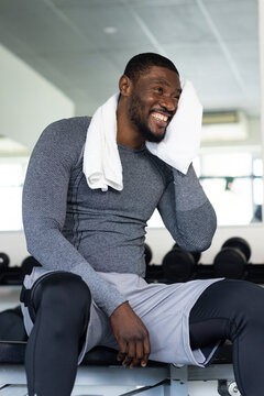 Mid adult man sitting on bench wearing gray compression top wiping sweat with towel at gym