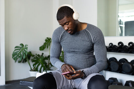African American mid-adult man sitting on gym bench checking pink smartphone, wearing headphones