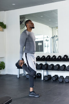 Mid adult African American man stretching quads in gym by dumbbell rack, wearing compression top