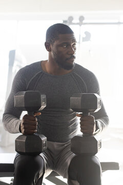 Man wearing grey long-sleeve top, leggings sitting on bench holding hex dumbbells in gym by windows