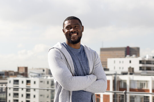 Mid-adult man standing on rooftop near metal railing, wearing grey hoodie and blue t-shirt, smiling