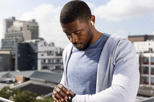 Mid-adult African American man checking smartwatch on rooftop, wearing grey hoodie and white earbud