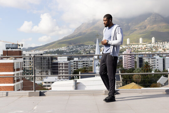 African American man standing on roof checking watch in hoodie earbuds shoes by railing, copy space