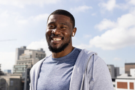 African mid-adult man standing and smiling on city rooftop, wearing light-gray hoodie, white earbud