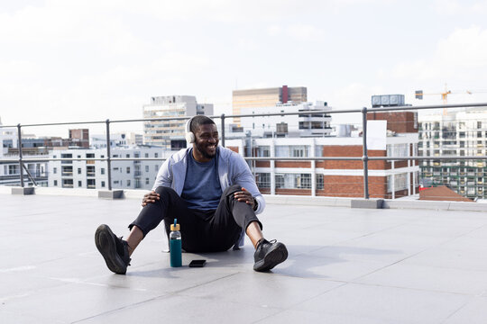 African American man sitting on roof in sportswear, wearing headset, bottle phone watch by railing